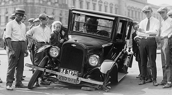 A black and white picture of an old automobile that has crashed on the side of the road with people standing around it.