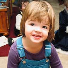 A little girl with short hair and bangs smiles at the camera. She is wearing a purple shirt and blue overalls.