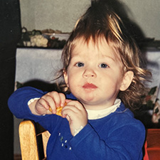 A young girl wearing a blue sweater eats a cracker. Her hair is in a ponytail on top of her head.