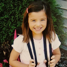 A young girl in a light shirt with brown hair and a backpack smiles at camera