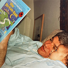An adult man lays in bed, holding a book. He is showing the book to a baby, who looks on with wide eyes.