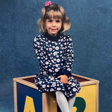 A young girl wearing a floral dress and tights sits on top of a large alphabet block. She is smiling, and her hair is tied up in a pink scrunchie.