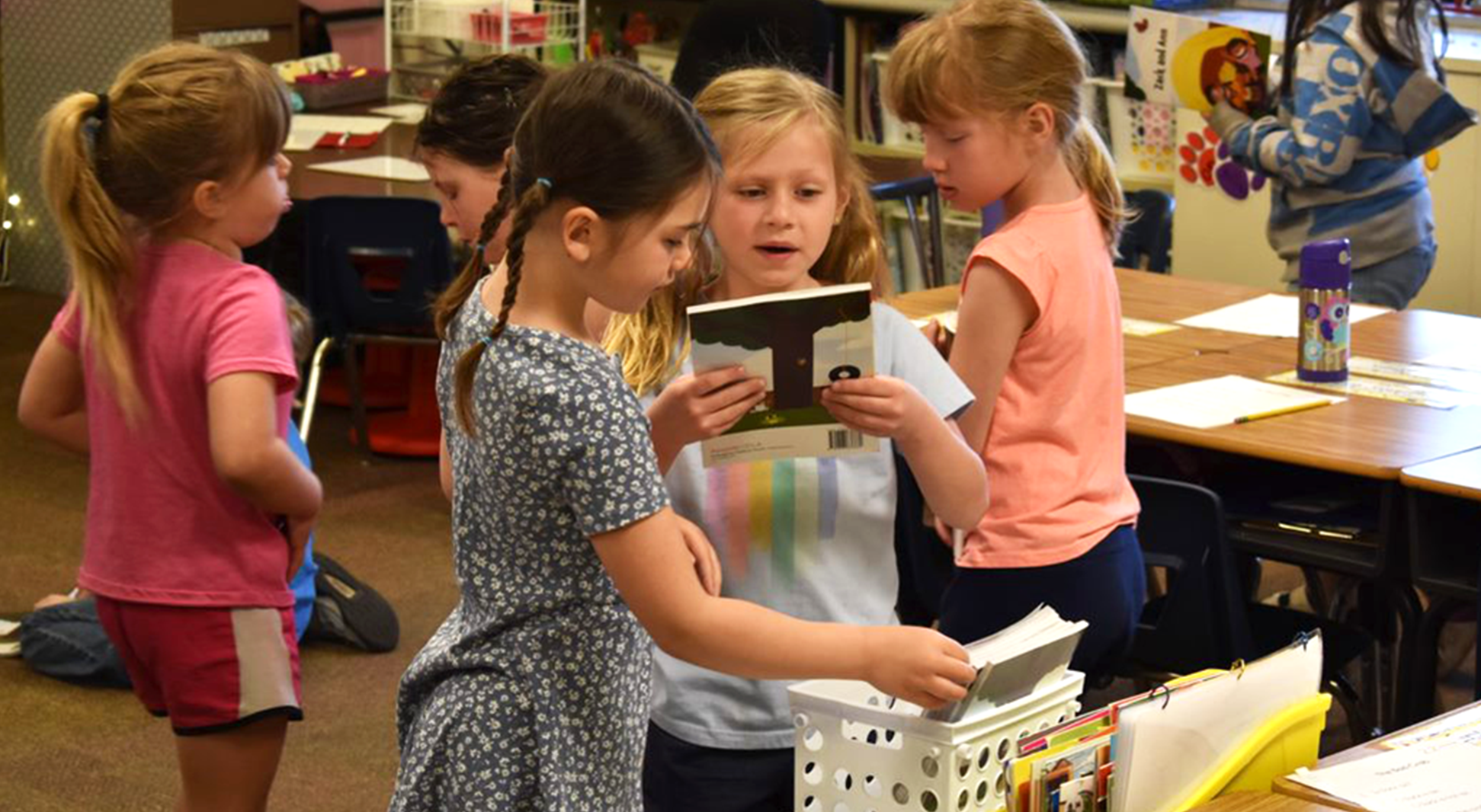 Two kindergarten students select picture books from a white crate. Other students are present in the background. Collins wears a blue dress and has dark brown braids.