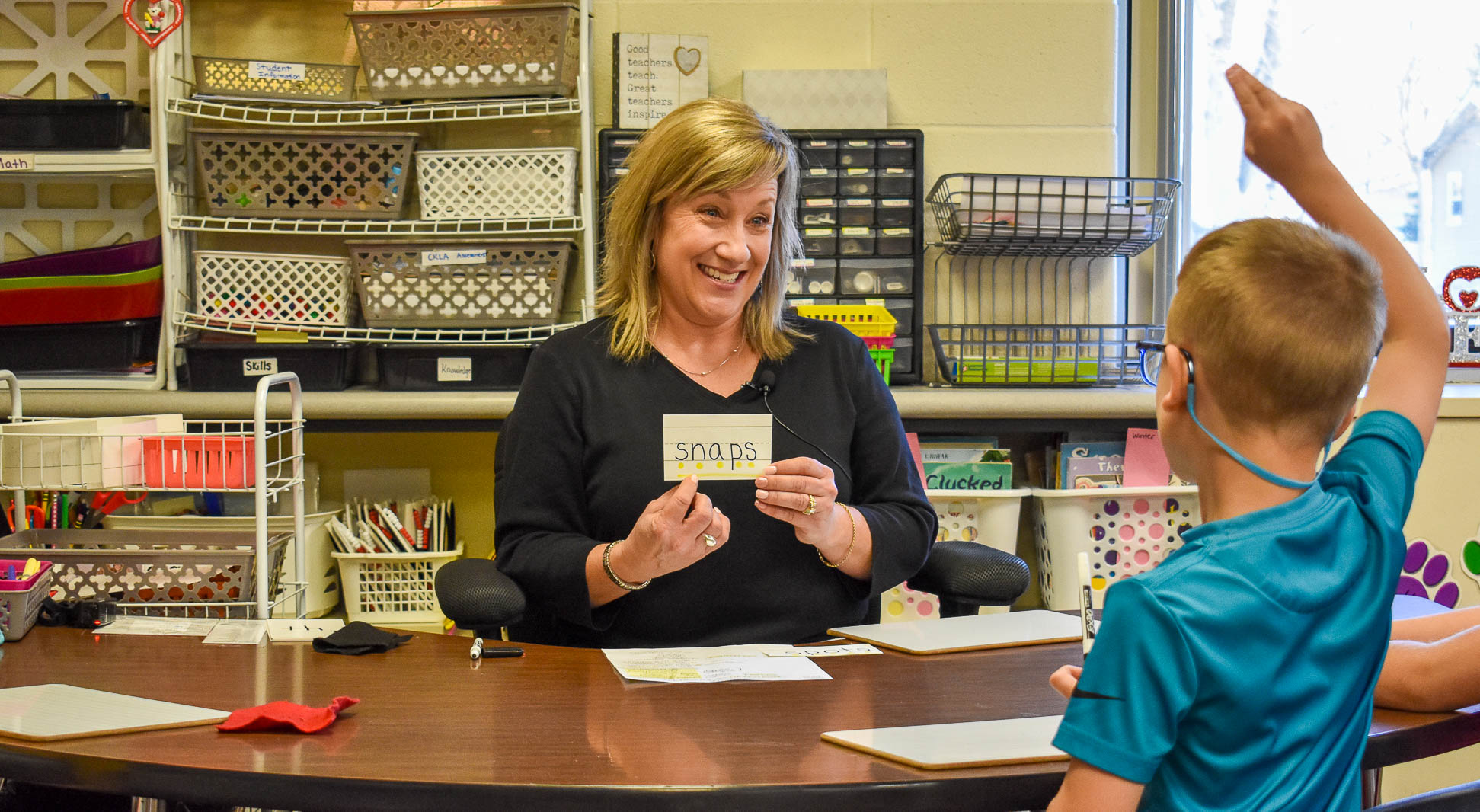 Tara Rabenberg, a teacher with shoulder-length blond hair and bangs, holds up an index card reading "snaps." She points at one of the letters and smiles at a young studnet in a blue shirt, who is sitting across from her and raising his hand.