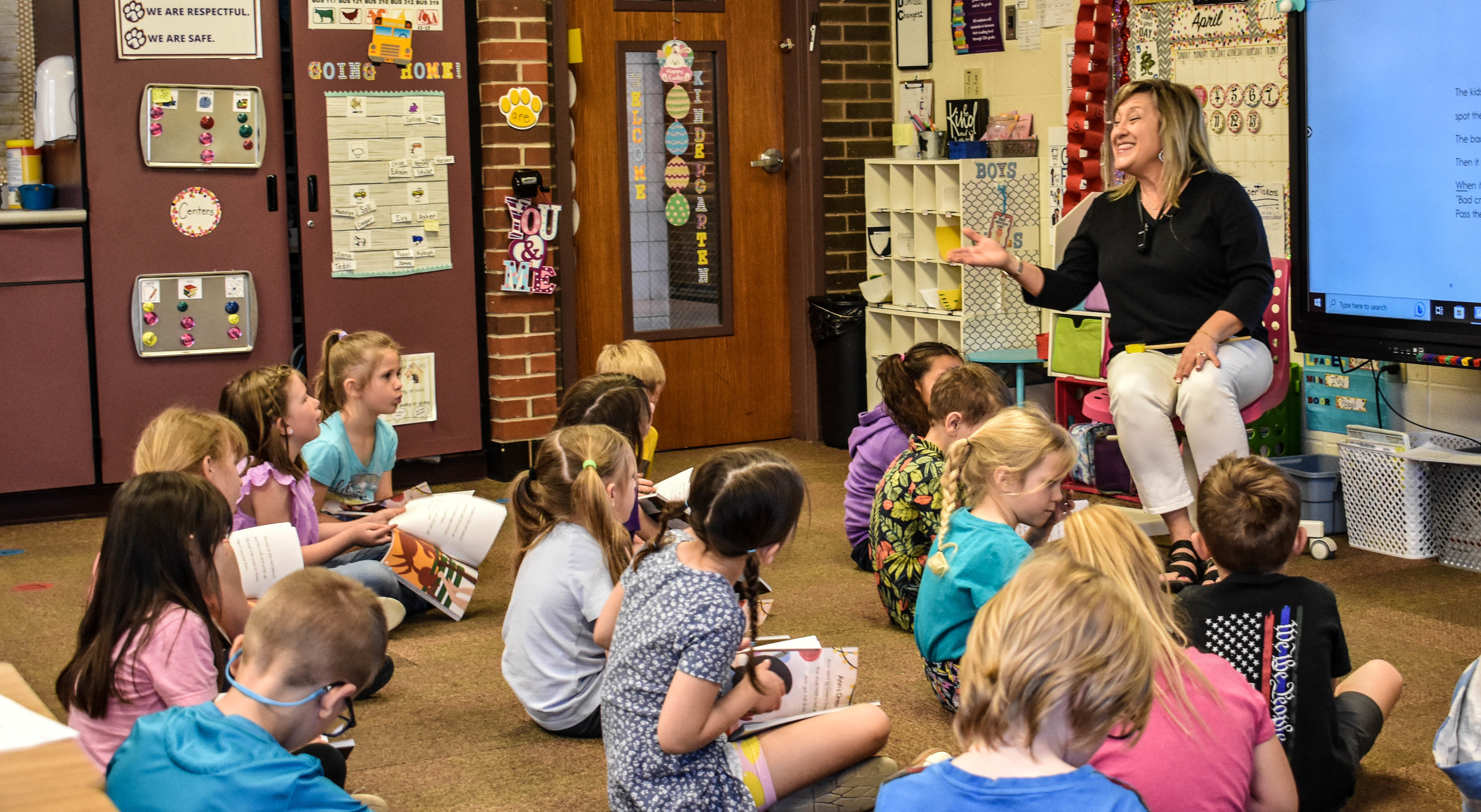 Teacher Tara Rabenberg sits in front of a class of kindergarteners, all of whom are seated on the floor and holding thin books. The text of the book is projected behind Rabenberg, and the rest of the room is covered with colorful posters.