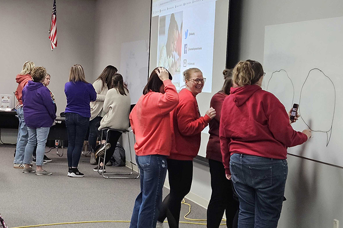 Three teachers draw on a white board while another group of teachers stands behind