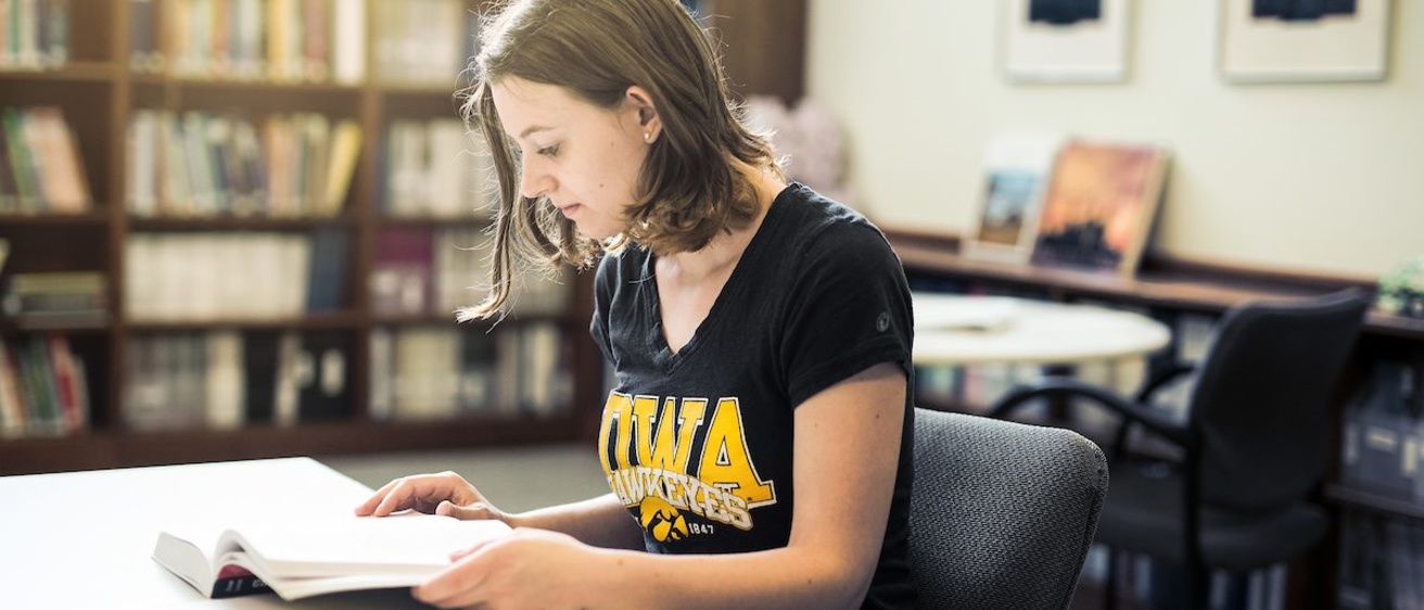 An Iowa student reads a book at a desk with bookshelves behind