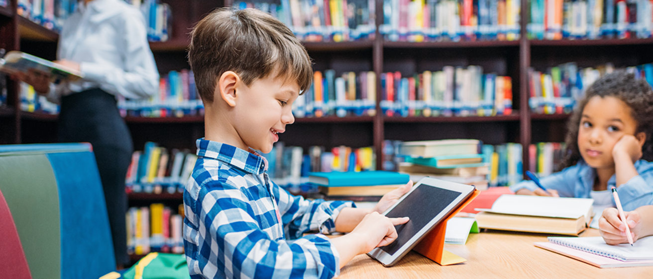 Young boy with brown hair uses a tablet to read at a library table