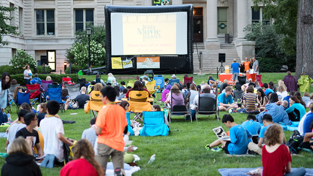 The lawn of the Pentacrest at the University of Iowa with a projector set up and people watching a movie in the grass