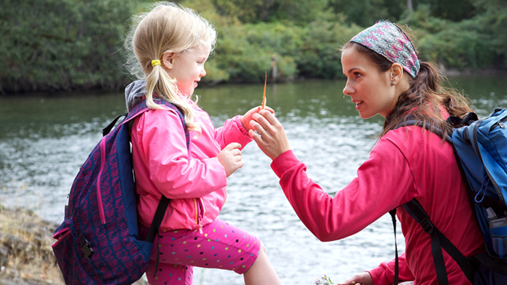 A mom and daughter, next to a pond, hold up a leaf to inspect it closer during a hike in nature.