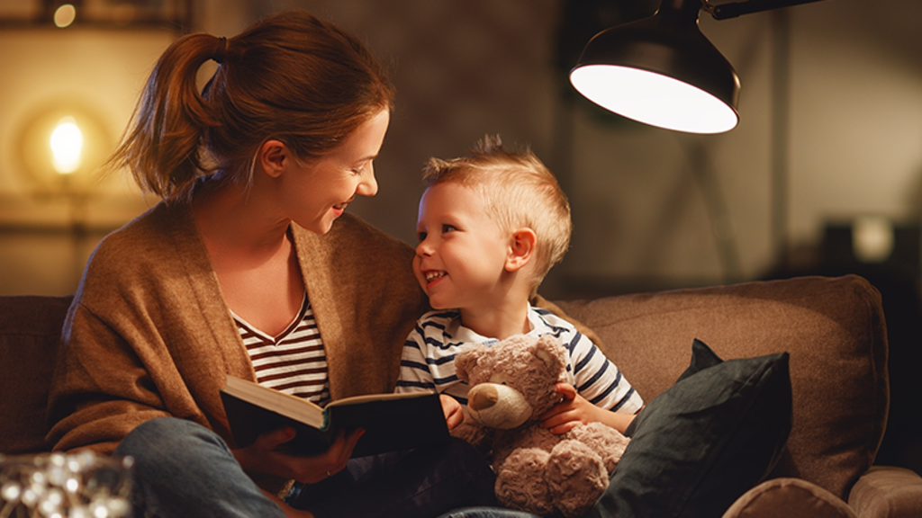 Mother and son sitting on a couch reading together under a lamp light with a teddy bear