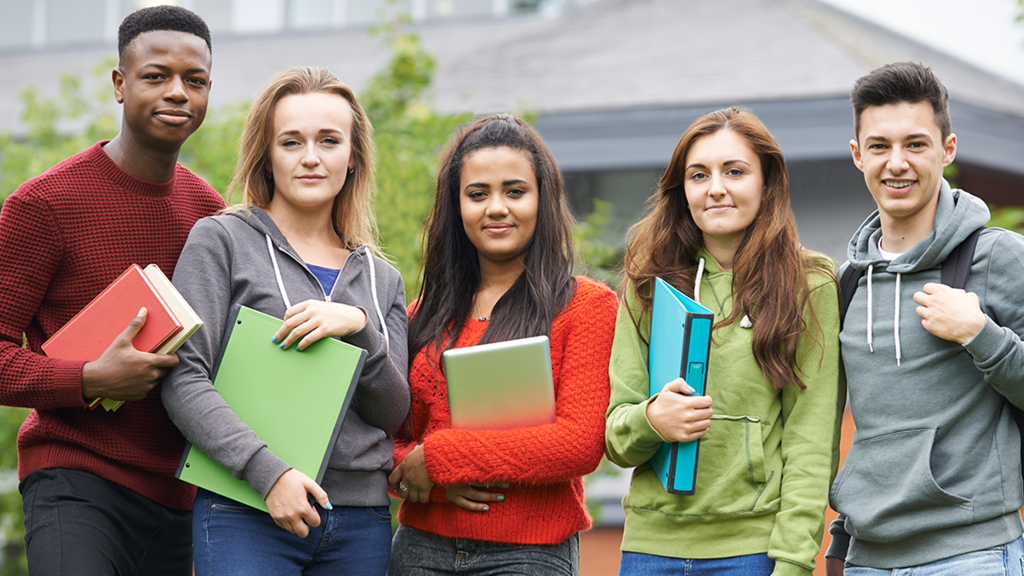 5 teens at school smiling for the camera with books, folders, or binders in their hands.